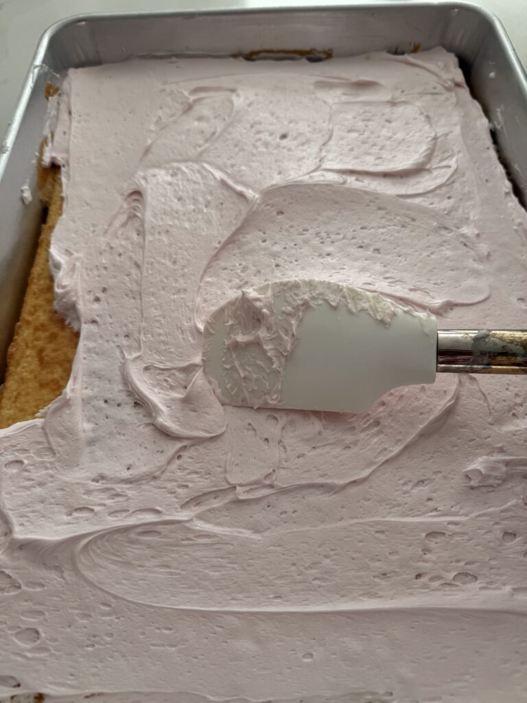 The back of a spatula spreads the pink icing onto the cake in a silver 18x13-inch cake pan on the counter.