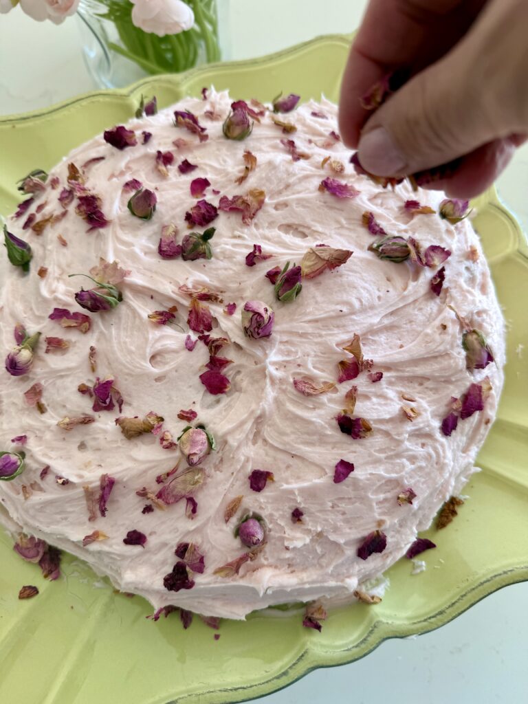 A finished Blush Buttercream Vanilla Cake on a light green cake stand is garnished with edible rose petals, with blush baby roses on the side of the counter.