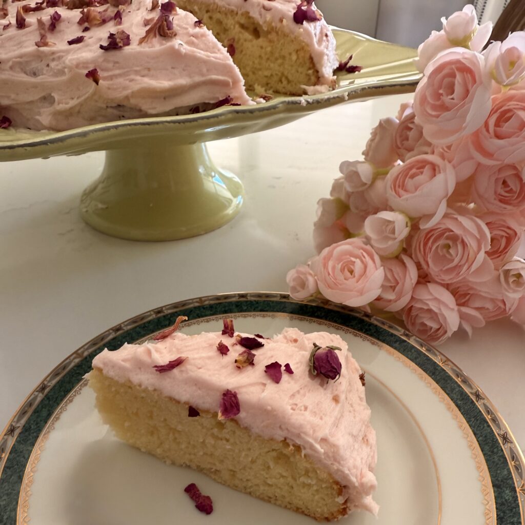 Blush Buttercream Vanilla Cake on a plate on the counter, with blush roses beside it. The entire cake is in the background on a light green cake stand.