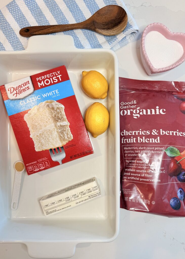 White baking dish with classic white cake mix, lemons, ground ginger in a teaspoon spoon, and butter on the counter. Beside the white dish is the bag of frozen mixed berries. Above is a pink heart dish with white sugar on the counter. Above the white dish is a blue and white dishcloth with a wooden spoon on the counter.