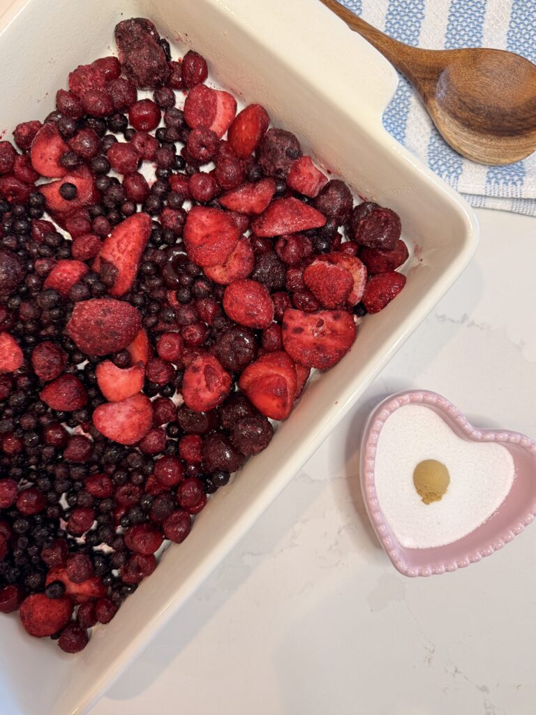 Frozen mixed berries in a 13x9 baking dish on the counter. With a pink heart-shaped bowl beside it with sugar and ground ginger in it on the counter. Above is a blue and white hand towel with a wooden spoon.