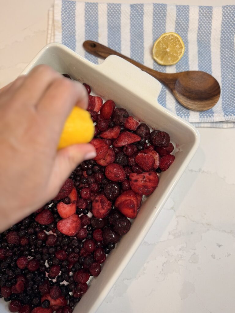 In a 13x9 baking dish are the frozen mixed berries with lemon juice on it on the counter, with a blue and white hand towel to eat top with a slice of lemon and a wooden spoon.