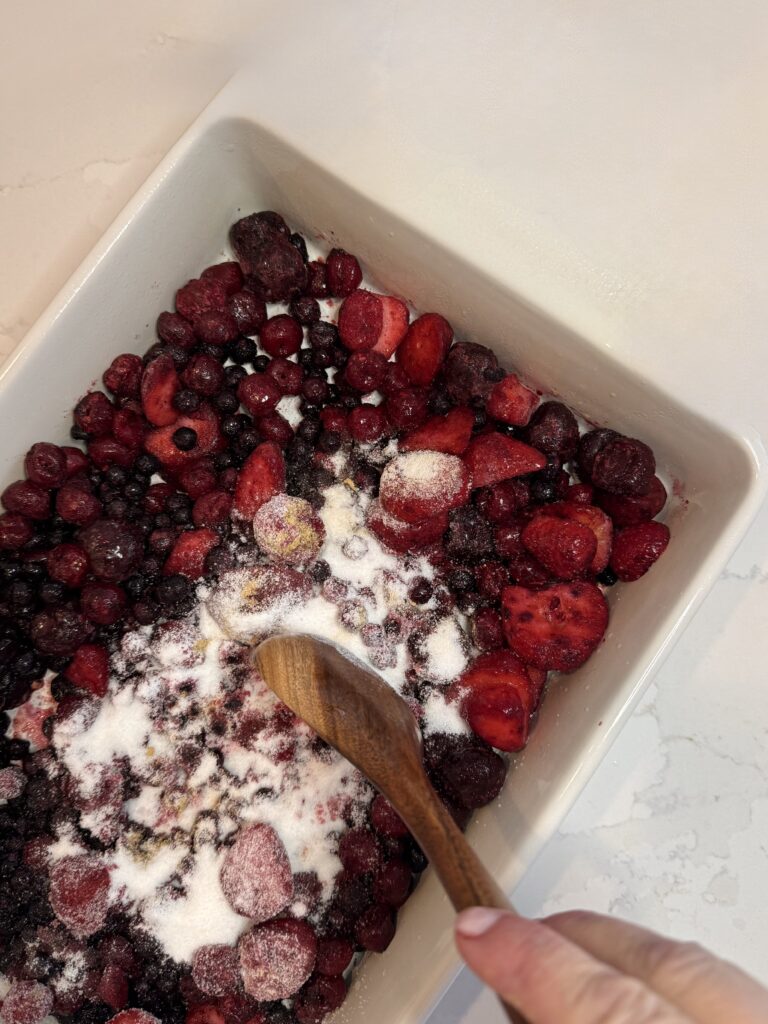 Mixing the sugar with the frozen berries with a wooden spoon in a white baking dish on the counter.