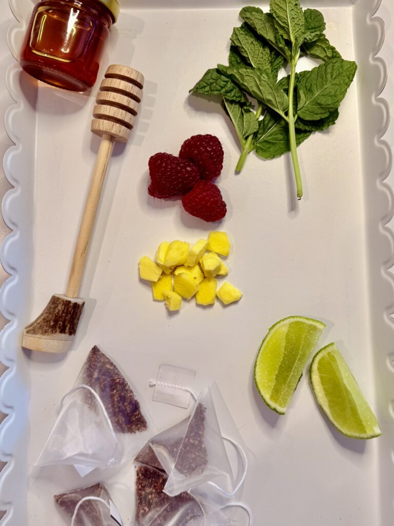 Caffeine-free raspberry tea bags, a piece of ginger, peeled and cut into pieces, a lime wedge for garnish, a jar of honey, mint leaves for garnish, and raspberries for garnish on a white tray on the counter.