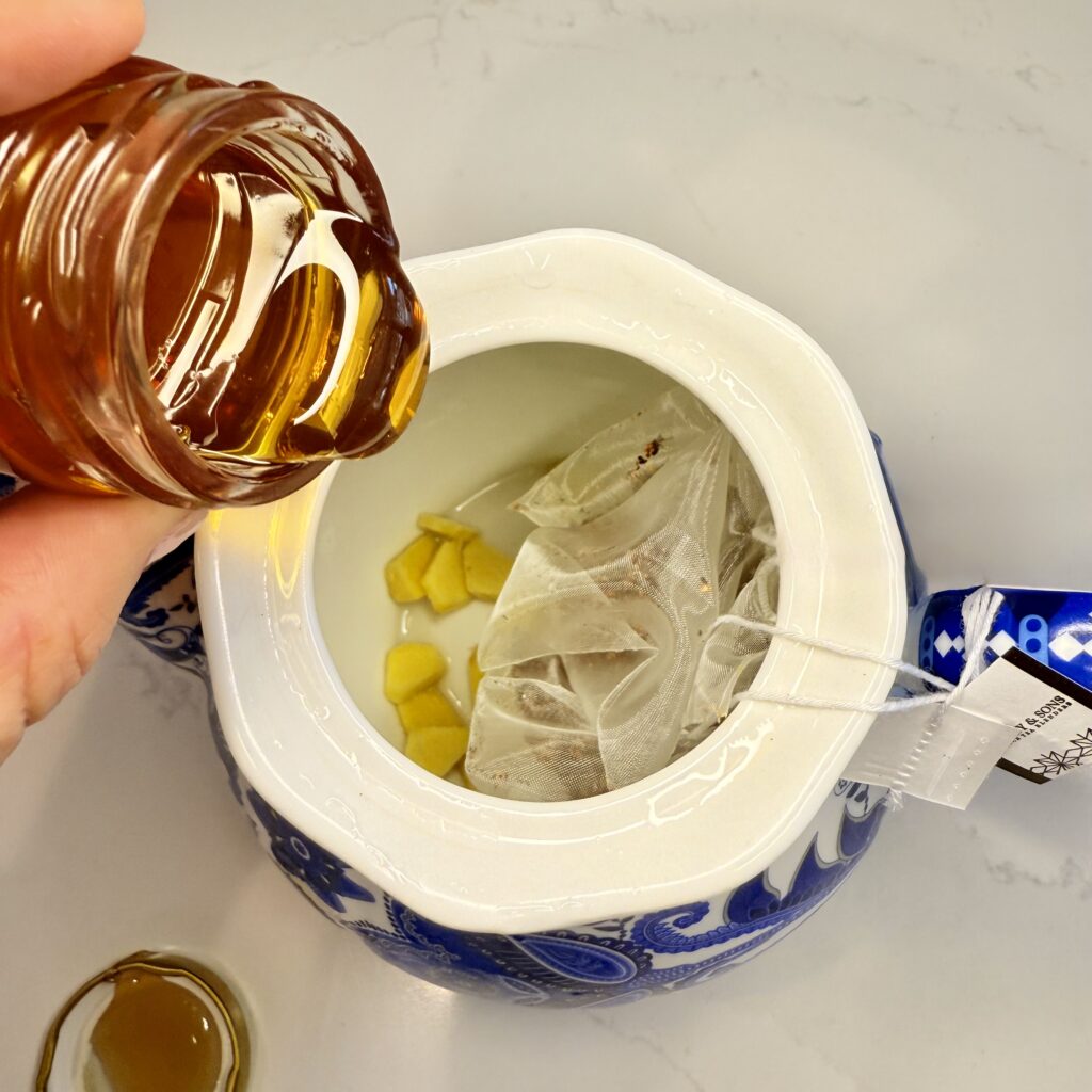 Pouring honey into a blue and white teapot with raspberry tea bags and pieces of ginger on the counter.