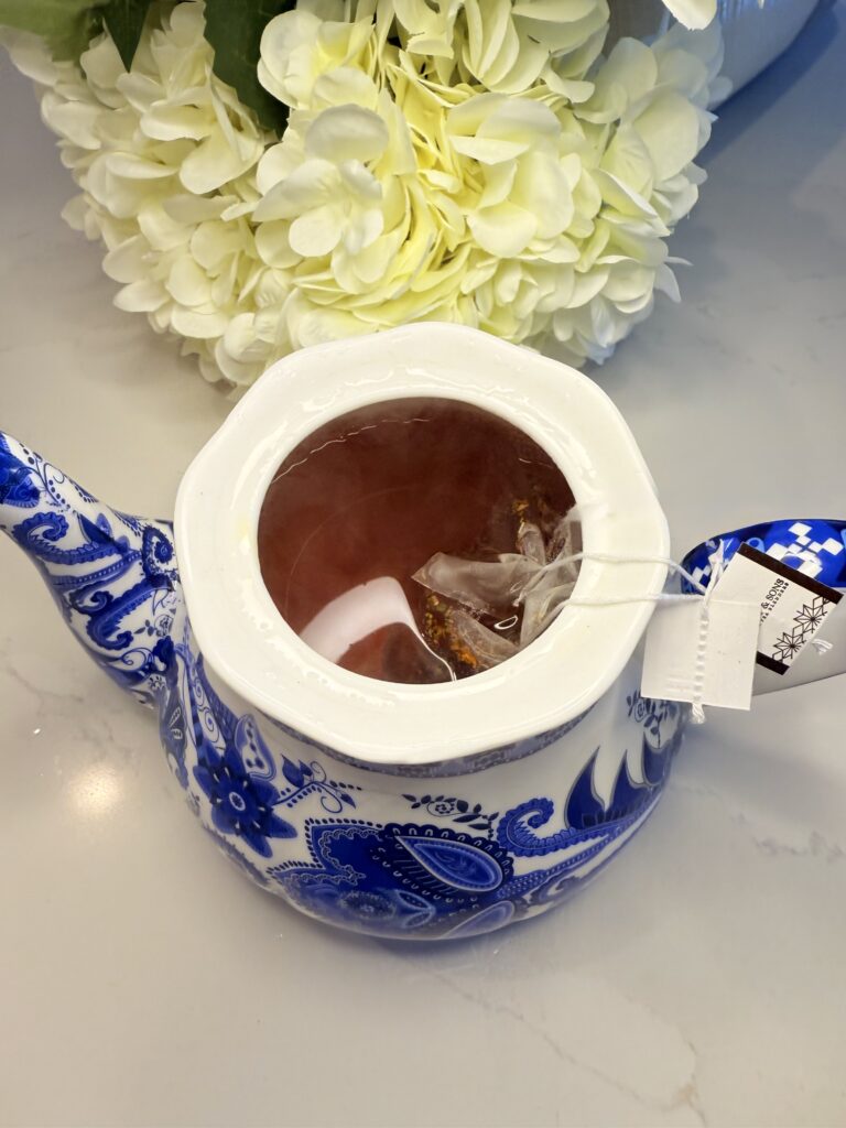 Blue and white teapot filled with honey, ginger pieces, and hot raspberry tea on the counter with fake white flowers in the background.