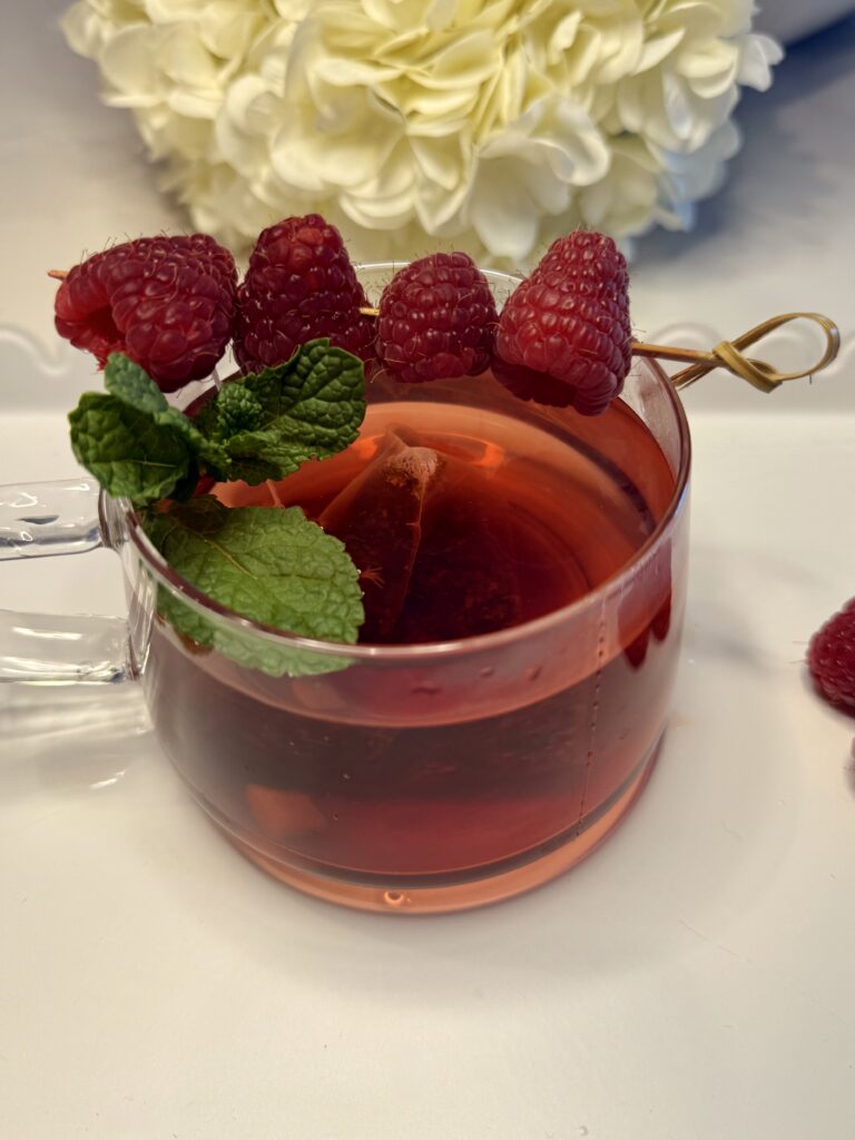 Warm raspberry ginger tea with mint leaves and fresh raspberries in a clear glass mug on the counter.