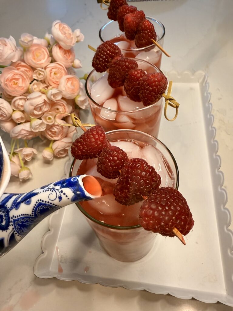 Pouring cooled raspberry tea into Three tall glasses of Raspberry Ginger Iced Tea garnished with raspberries on the counter.