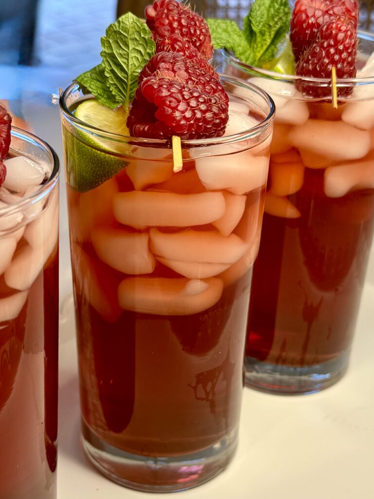 Three tall glasses of Raspberry Ginger Iced Tea garnished with raspberries and mint leaves on the counter.