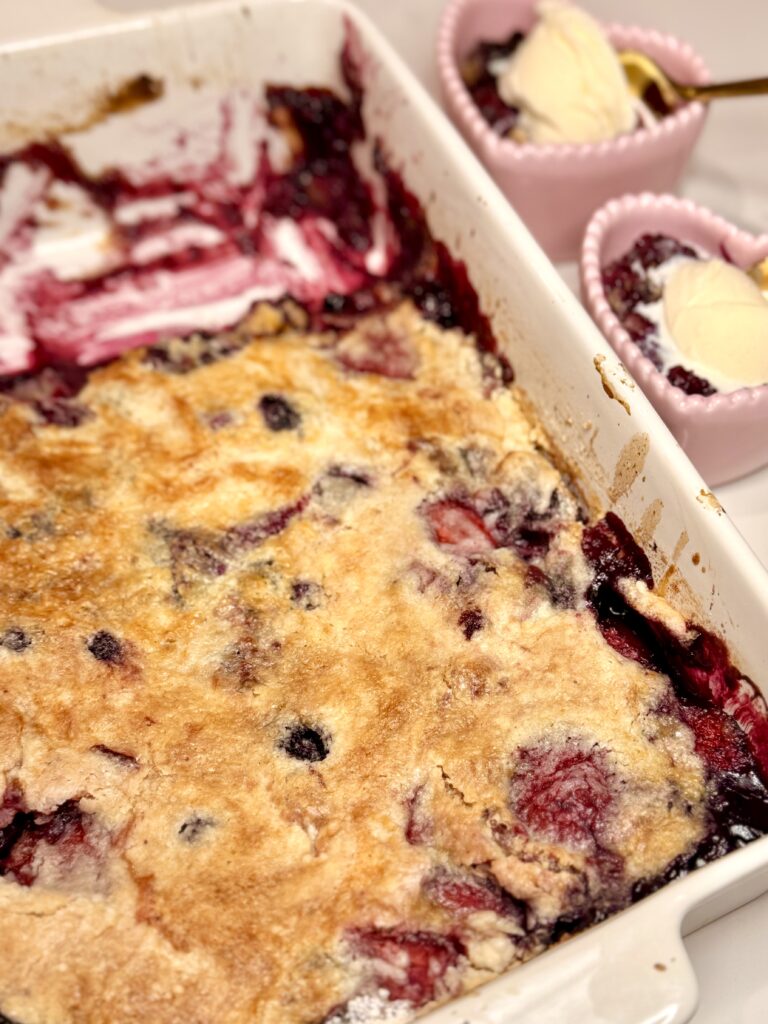 Easy Berry Cobbler is in a white dish with pink heart bowls filled with cobbler and vanilla ice cream on the the side of the cobbler white baking dish on the counter. 