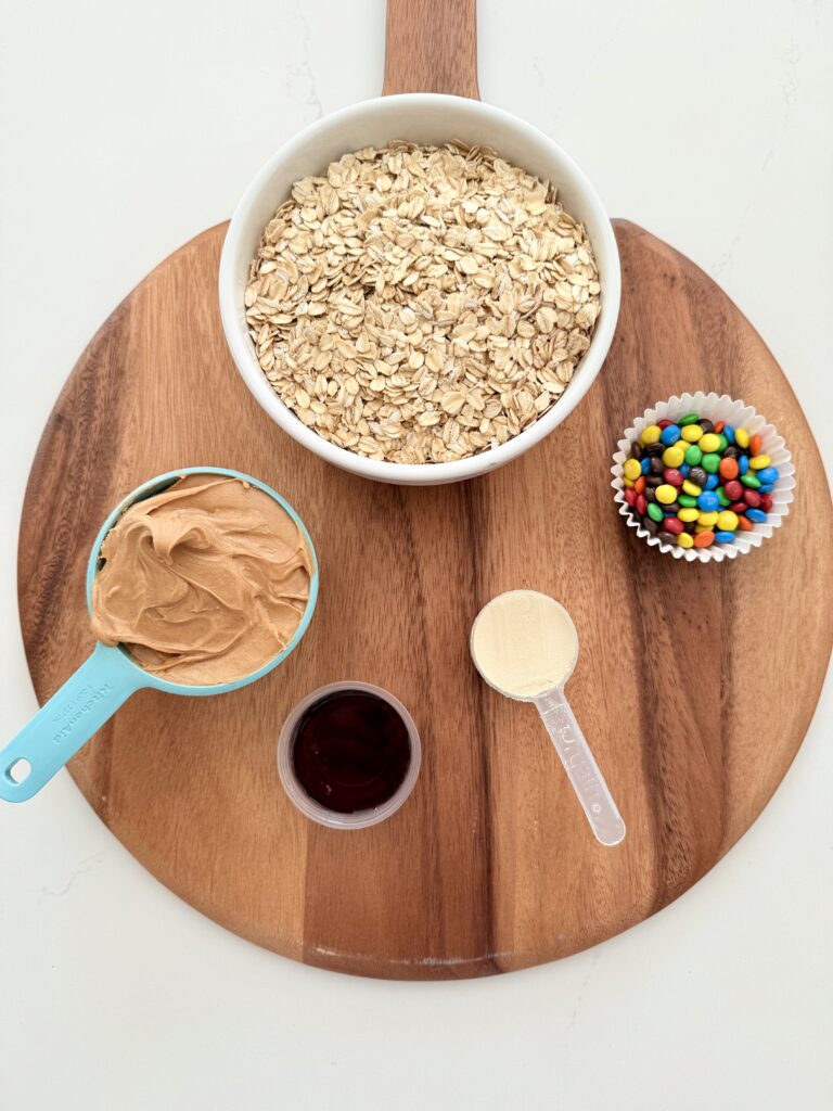 Protein powder, mini M&M candies in a paper muffin cup, peanut butter in a measuring cup, maple syrup in a mini plastic cup, and rolled coated in a large white mixing bowl on a brown cutting board on the counter.