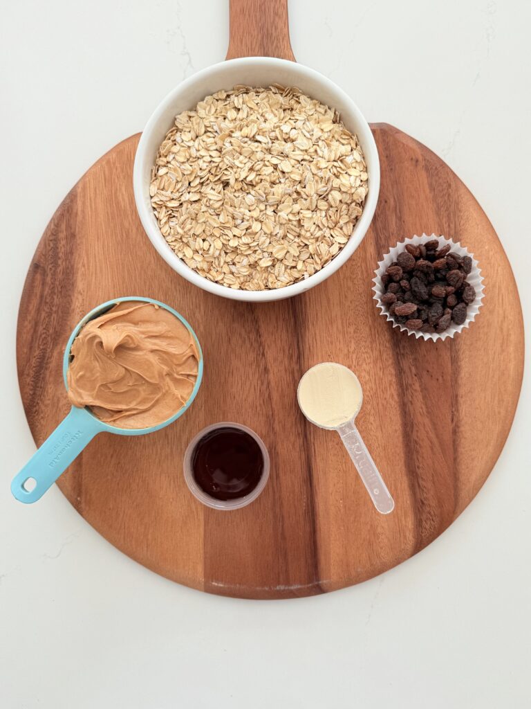 Rolled oats, peanut butter, raisins, protein powder, and maple syrup in individual containers on a brown cutting board on the counter.