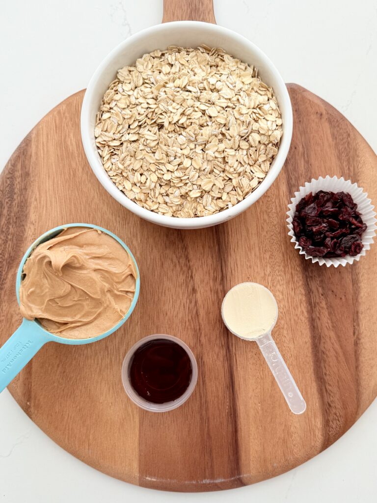 The protein powder, peanut butter, dried cranberries, maple syrup and the rolled oats are in individual containers and placed on a brown cutting board on the white counter.