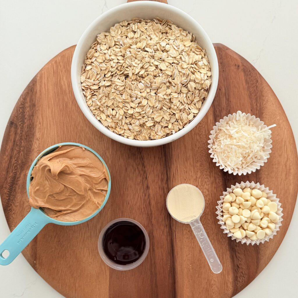 Rolled oats in a white bowl, peanut butter in a blue measuring cup, maple syrup in a plastic container, protein powder in a measuring cup, white chocolate chip in a muffin cup, and coconut flake in a muffin cup on a brown cutting board on the counter.