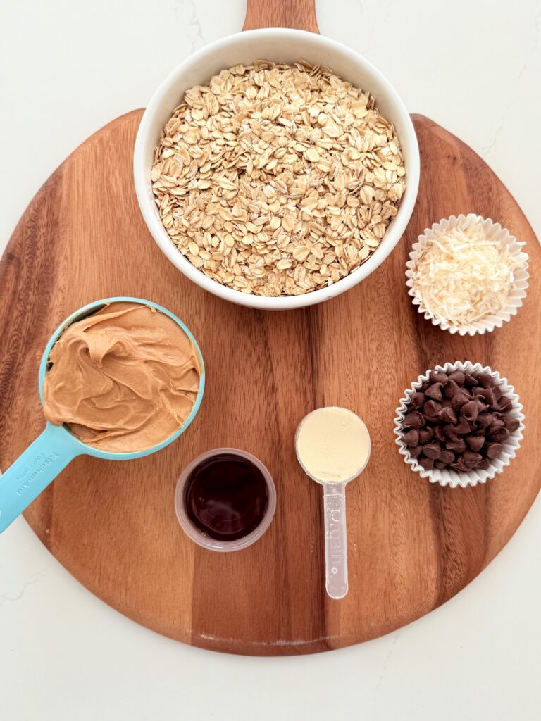 Vanilla protein powder, rolled coated, keto syrup, peanut butter semi-sweet chocolate chips, and coconut flakes in separate containers on the counter.