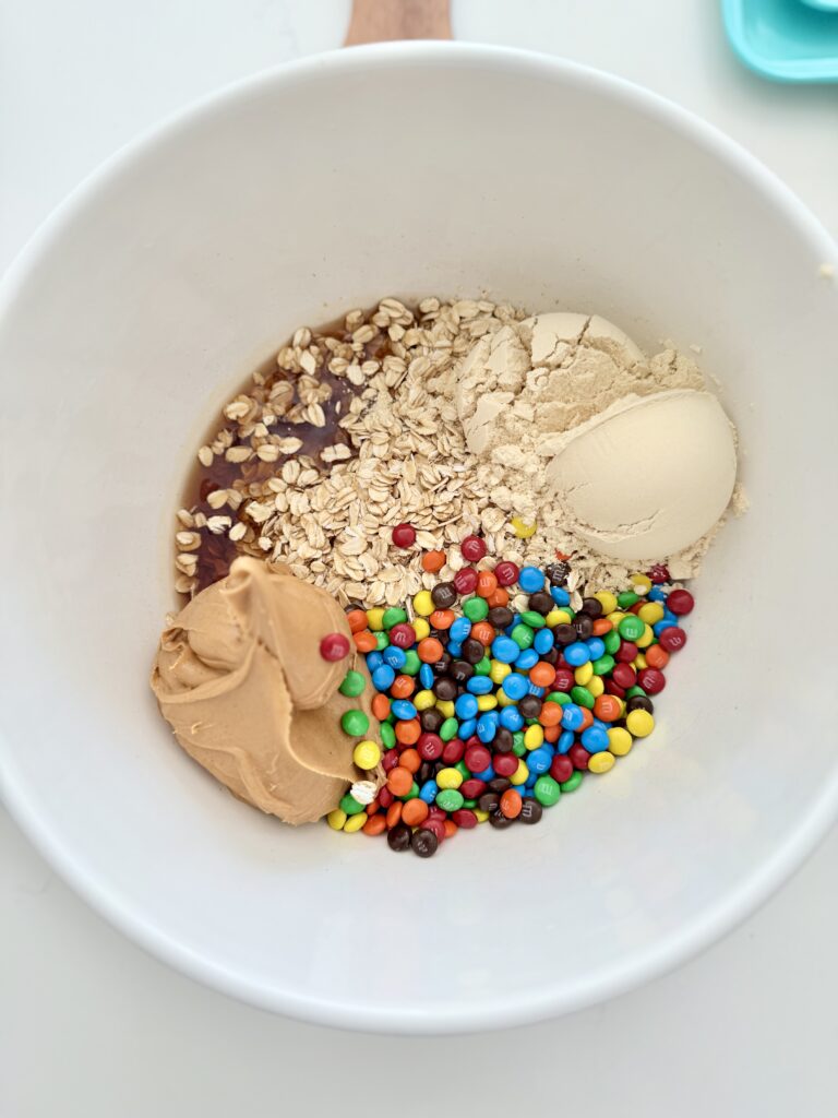 Protein powder, mini M&M candies, peanut butter, maple syrup and rolled coated in a large white mixing bowl on the counter.