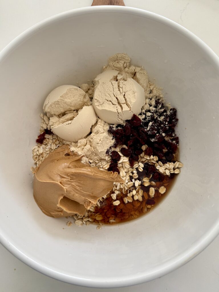 Protein powder, peanut butter dried cranberries, maple syrup and oats in a large white mixing bowl on the counter.