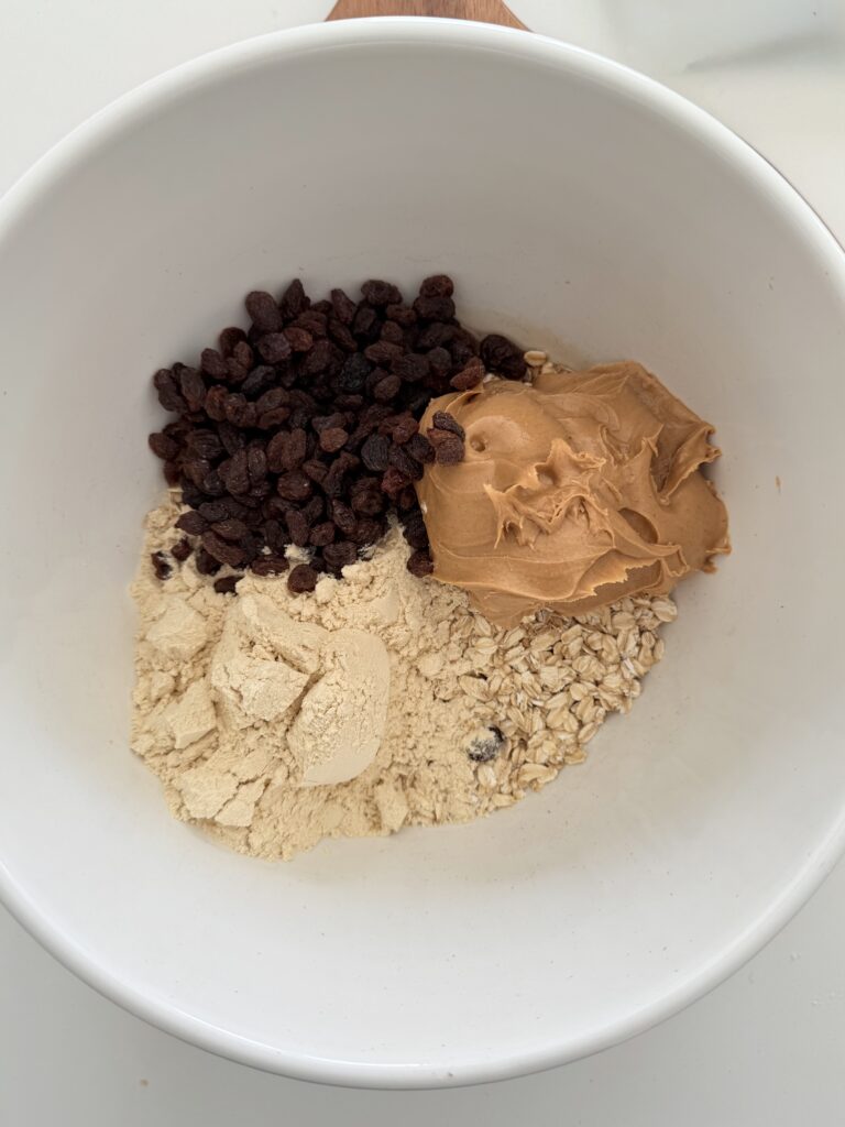 raisins, peanut butter, oats, maple syrup and protein powder in a large mixing bowl on a brown cutting board.