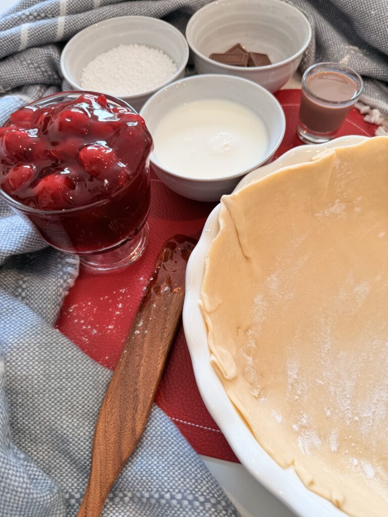 Cherry pie filling in a glass, milk in a bowl, pearl sugar in a bowl, chocolate pieces in a bowl, chocolate liqueur in a shot glass and pie crust fit into a 10 inch pie pan on a red mat on the counter. Also a blue blanket surrounds the ingredients on the counter.