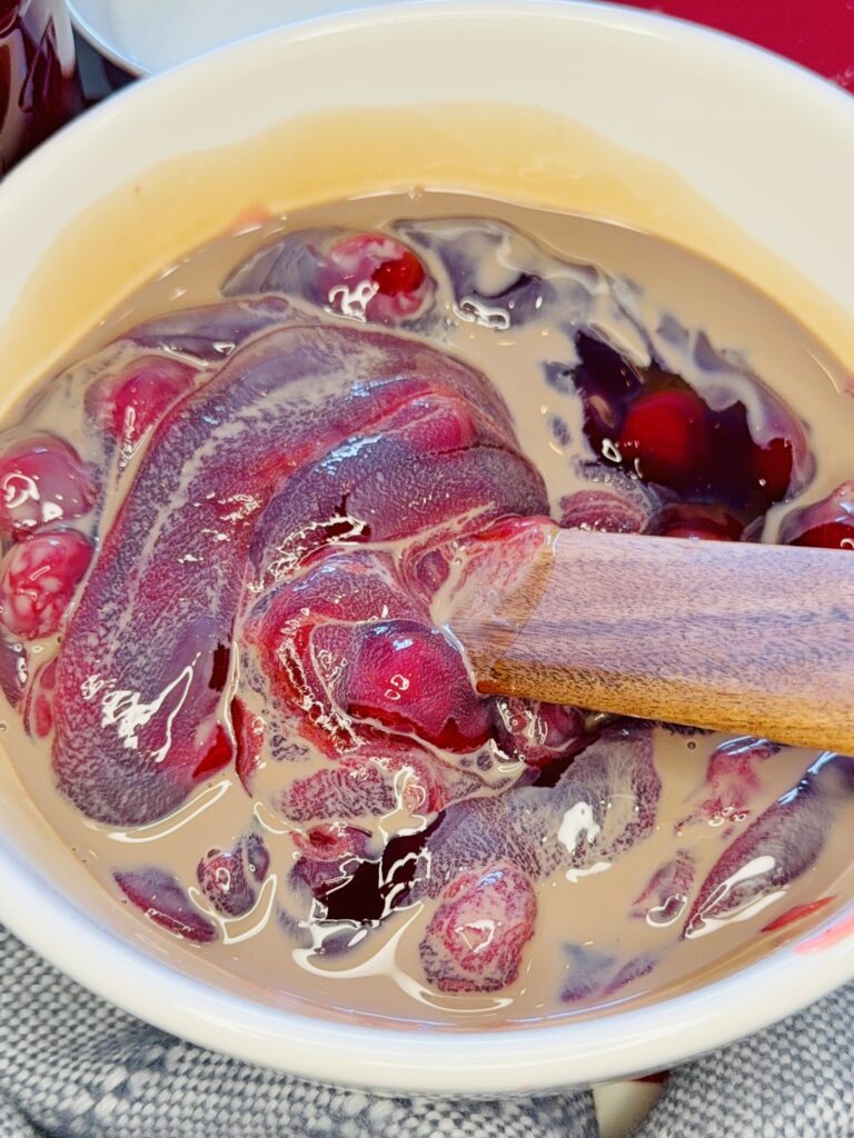 A wooden spoon storing the chocolate liqueur with the canned cherry pie fill in a white mixing bowl on the counter.