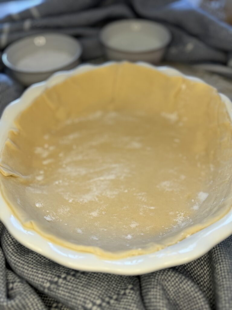 Pie crust in a 10 inch pie plate on the counter surrounded by a blue blanket and two small white bowls.