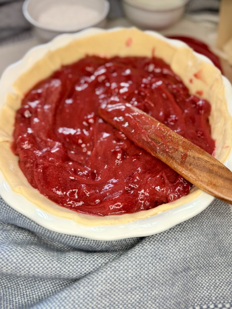 A wooden spoon smoothing out the pie filling in an unbaked pie crust on a white pie plate on the counter.