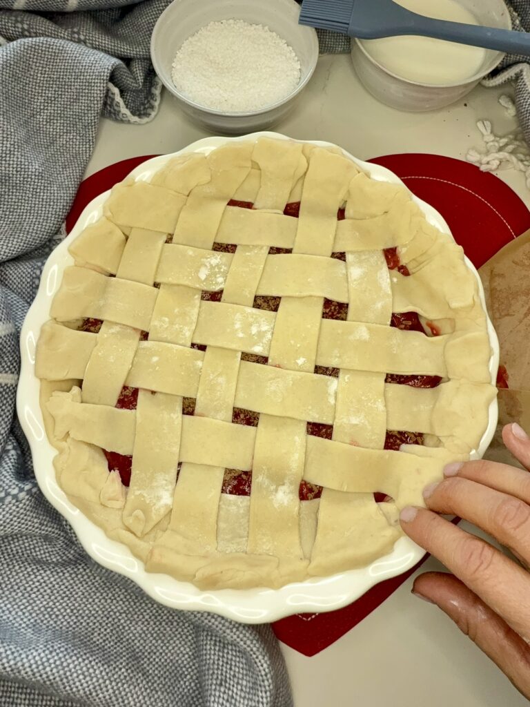 Weaved long pieces of raw pie crust, making a lattice pattern on top of the pie filling on the counter. Pressing and sealing the pie edges with fingers.