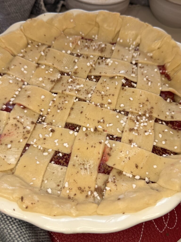 Sugar and grated chocolate on top of the weaved long pieces of raw pie crust, making a lattice pattern on top of the pie filling on the counter.