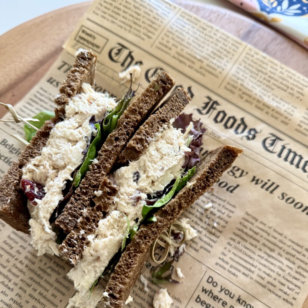 chicken salad as a sandwich with brown bread on newspaper parchment paper on a brown cutting board on the counter.