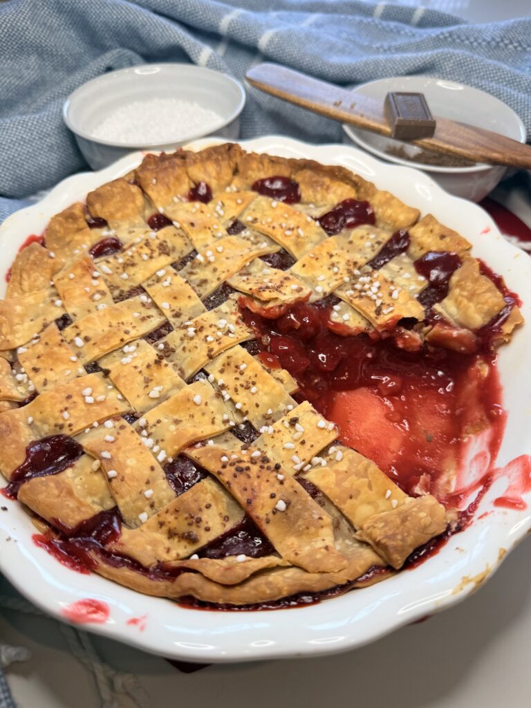 Chocolate Cherry Pie missing a piece in a white pie plate with sugar and chocolate in white bowls surrounded by a blue rag in the back ground on the counter.