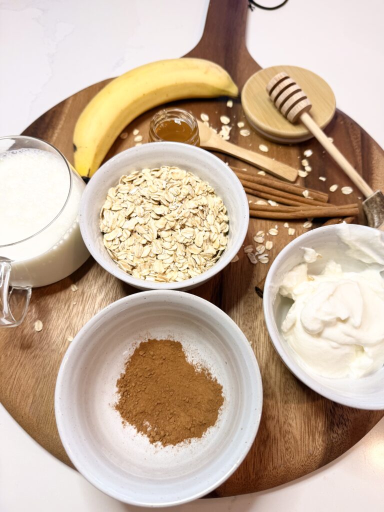 Rolled oats, a banana, vanilla yogurt, milk, cinnamon, and honey and honey dipper, and cinnamon sticks are on a brown cutting board that is on the counter.