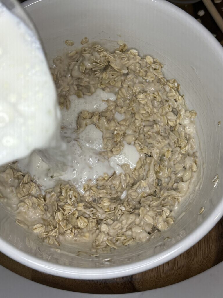  Banana and cinnamon overnight oats ingredients in a bowl, with milk being poured into the bowl, on a brown cutting board.