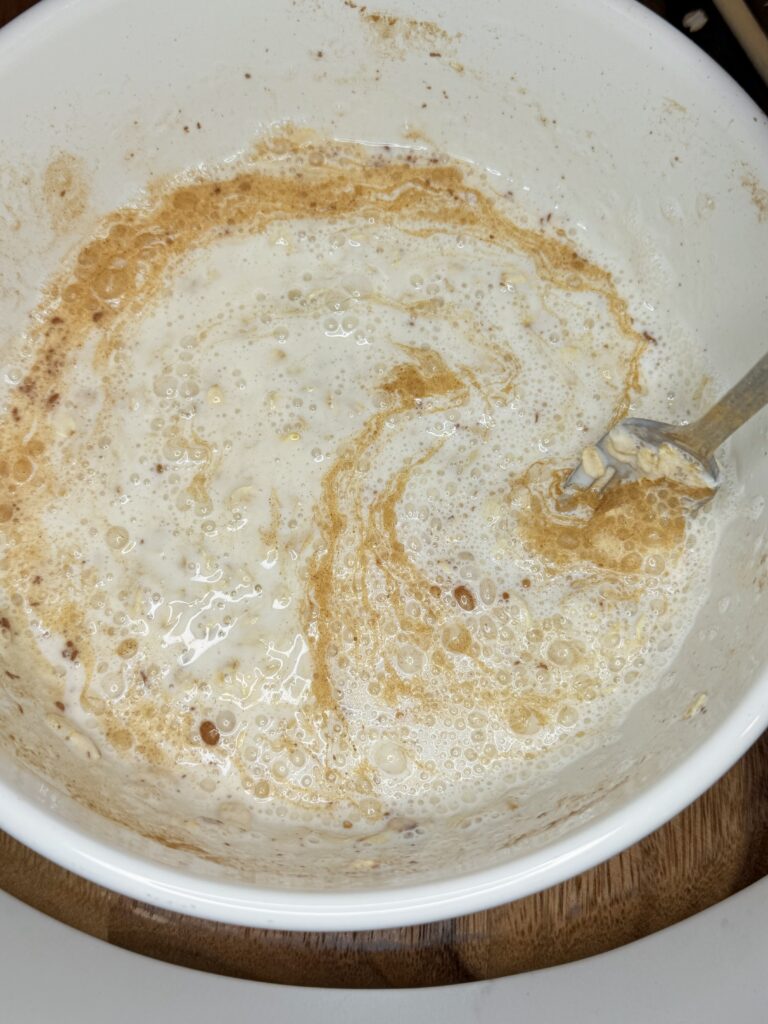 Banana and cinnamon overnight oats in a bowl being stirred with a fork on a brown cutting board.