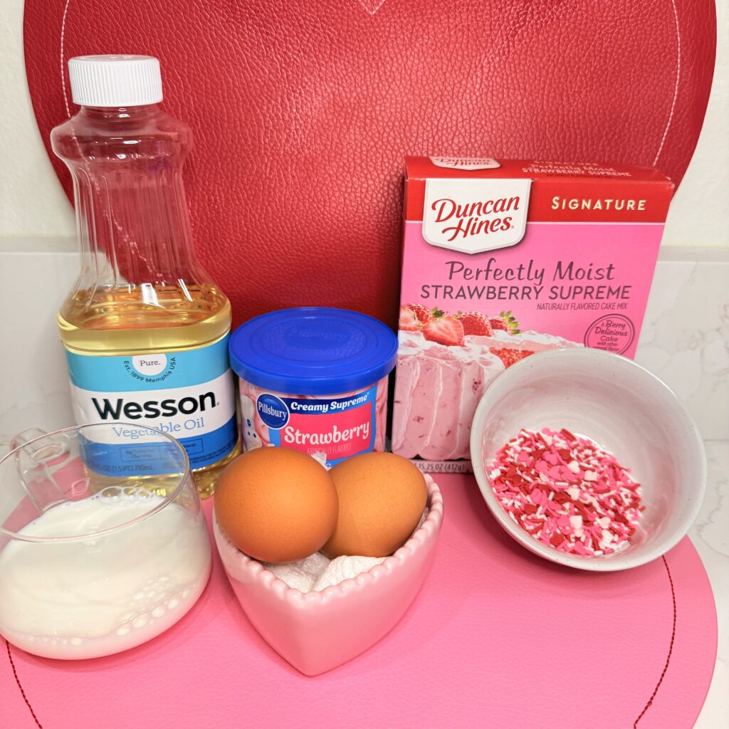 Strawberry cake mix, strawberry frosting, vegetable oil, milk, eggs and valentine themed sprinkles on a pink heart shaped placemat on the counter.