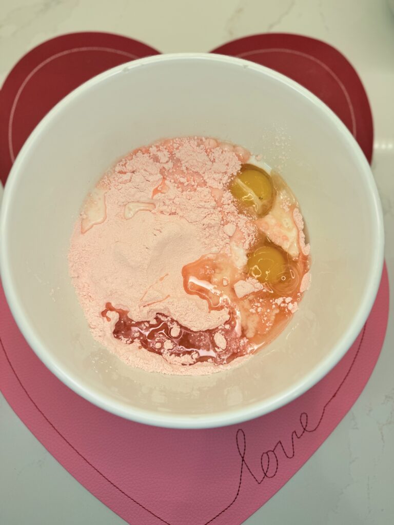 Egg, oil, milk, and strawberry cake mix in a large white mixing bowl on red and light pink placemats on the counter.