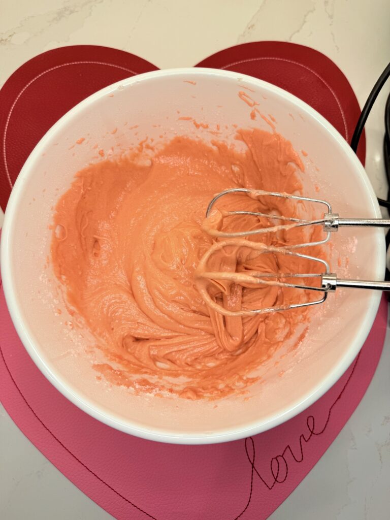 Mixed strawberry cake batter in a large white mixing bowl on red and light pink placemats on the counter.