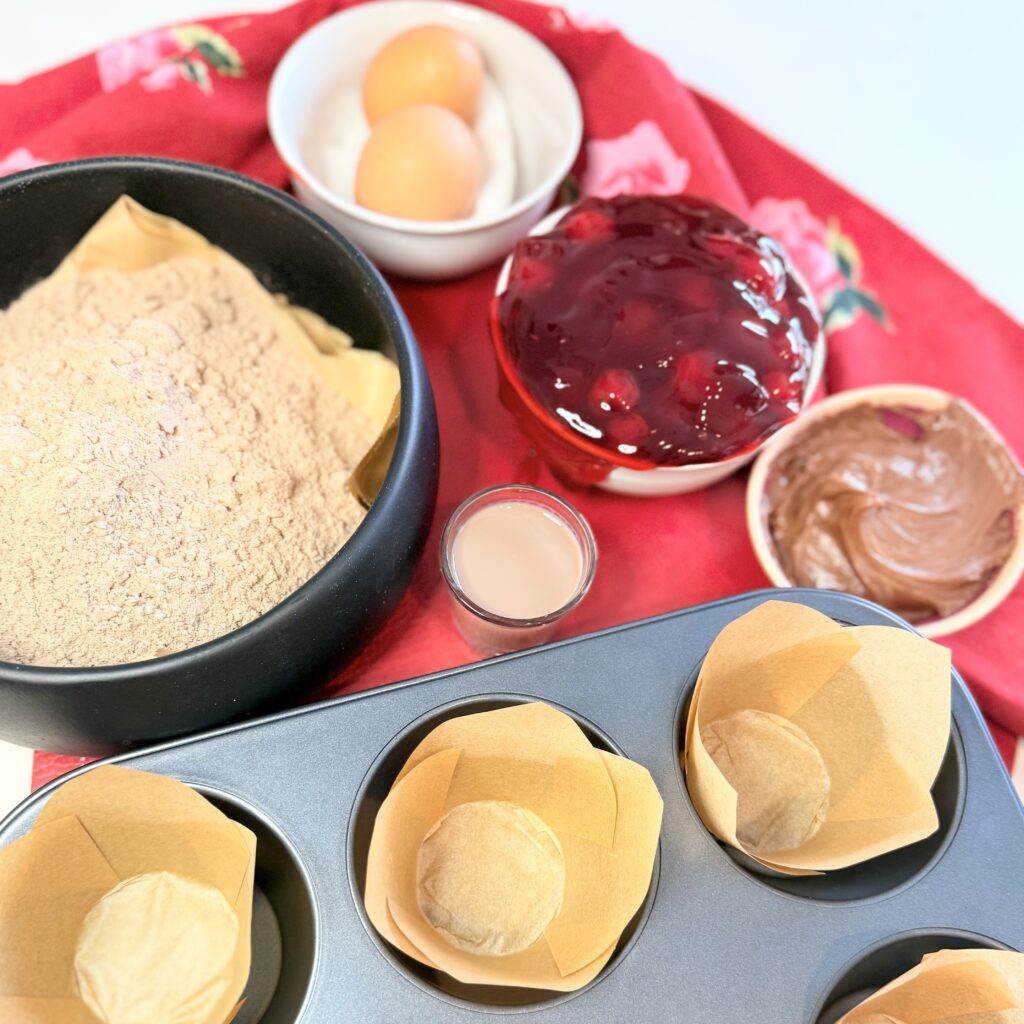 Eggs in a small bowl, cherry pie filling in a bowl, chocolate frosting in a small bowl, chocolate liqueur in a shot glass, chocolate cake mix in a black bowl and jumbo muffing liners in muffing cups on a red cloth on the counter.