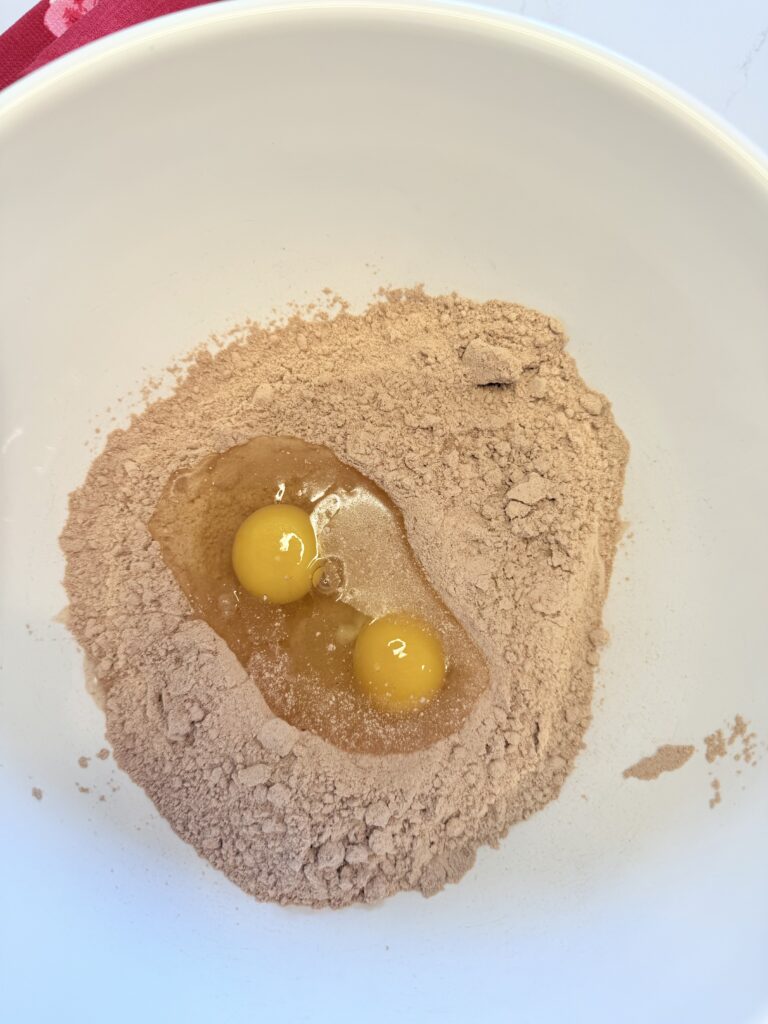 Two raw eggs on top of chocolate cake mix in a large white mixing bowl on the counter.