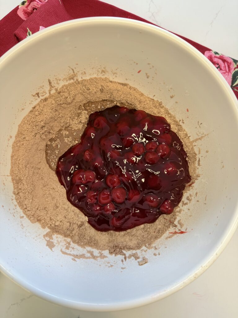 Cherry pie filling on top of mixed together eggs and cake batter in a large white mixing bowl on the counter.