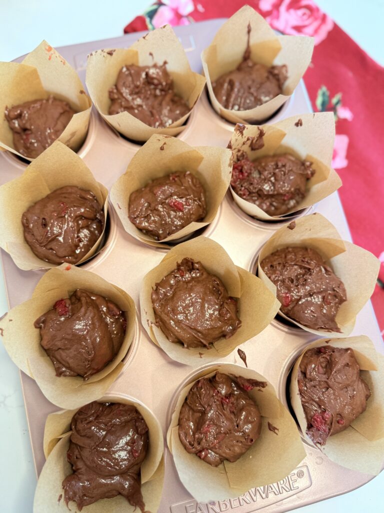 Chocolate cherry muffin raw batter in a pink muffin tin that is lined with jumbo muffin cups- on a red hand towel on the counter.