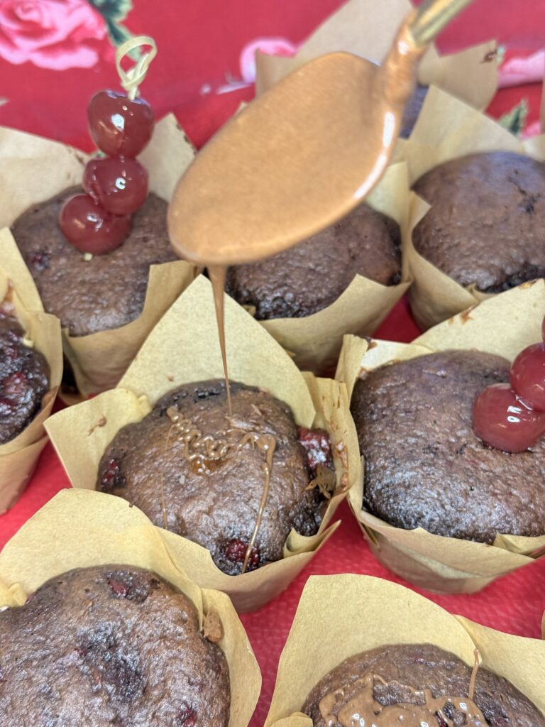 Drizzling warmed chocolate frosting on top of the muffins on a red hand towel on the counter.