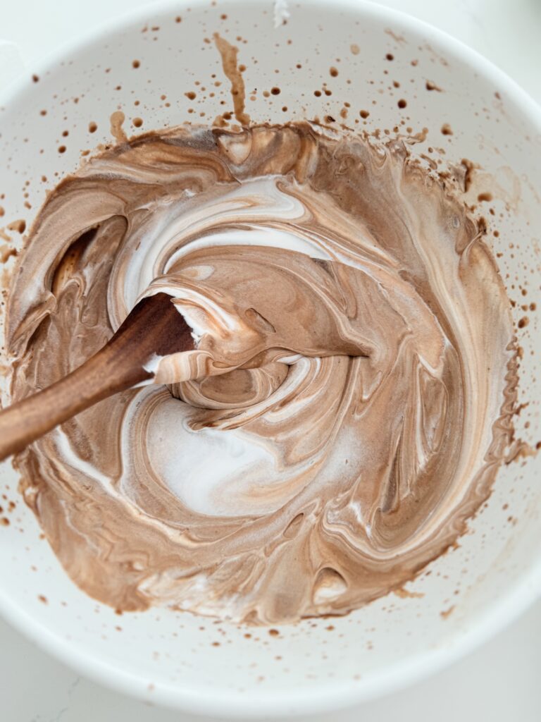 A wooden spoon mixing Cool Whip and chocolate pudding in a large mixing bowl on the counter. 