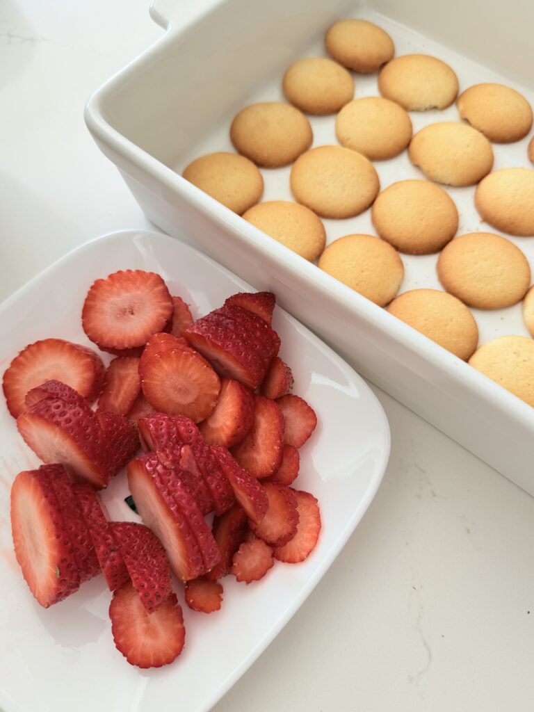 Sliced strawberries on a white plate beside vanilla wafers in a white baking dish on the counter.