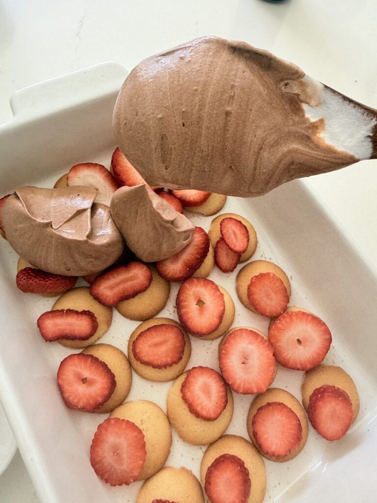 topping chocolate pudding and cool whip mixture on top of strawberries and vanilla wafer cookies in a white baking dish on the counter.