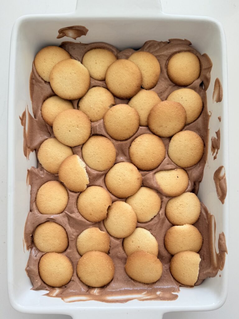 Vanilla wafer cookies layered on top of chocolate pudding and cool whip mixture in a white baking tray on the counter.