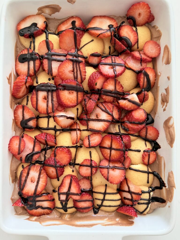 Layers of chocolate pudding, vanilla wafer cookies, and sliced strawberries, topped with dark chocolate sauce in a white baking tray on the counter.