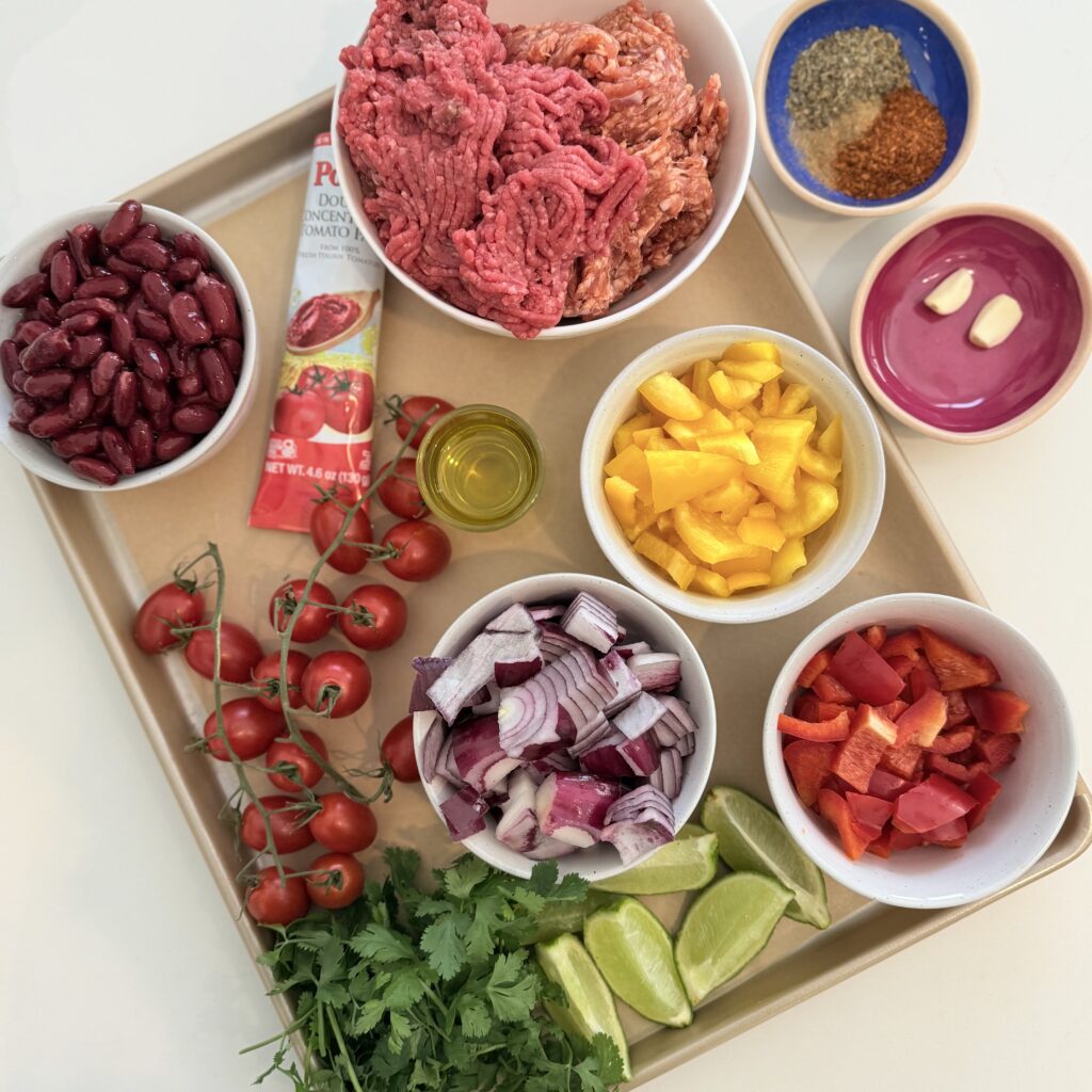 Olive oil, chopped red onion, garlic cloves, chopped red and yellow bell pepper, kidney beans, ground spicy pork sausage, ground beef, cajun seasoning, ground cumin, dried oregano, tomato paste in a tube, fresh cilantro, limes cut into wedges, and cherry tomatoes on a sheet pan on the counter.