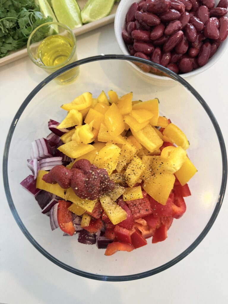 Olive oil, chopped red onion, garlic cloves, chopped red and yellow bell pepper, kidney beans, salt and pepper, and tomato paste in a glass bowl on the counter.