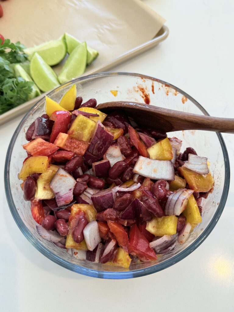 Olive oil, chopped red onion, garlic cloves, chopped red and yellow bell pepper, kidney beans, salt and pepper, and tomato paste in a glass bowl on the counter.