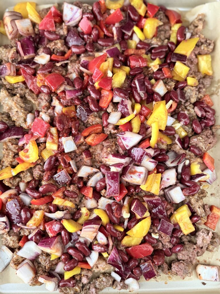 Olive oil, chopped red onion, garlic cloves, chopped red and yellow bell pepper, kidney beans, ground spicy pork sausage, ground beef, cajun seasoning, ground cumin, dried oregano, tomato paste in a tube, fresh cilantro, limes cut into wedges, and cherry tomatoes on a sheet pan on the counter.